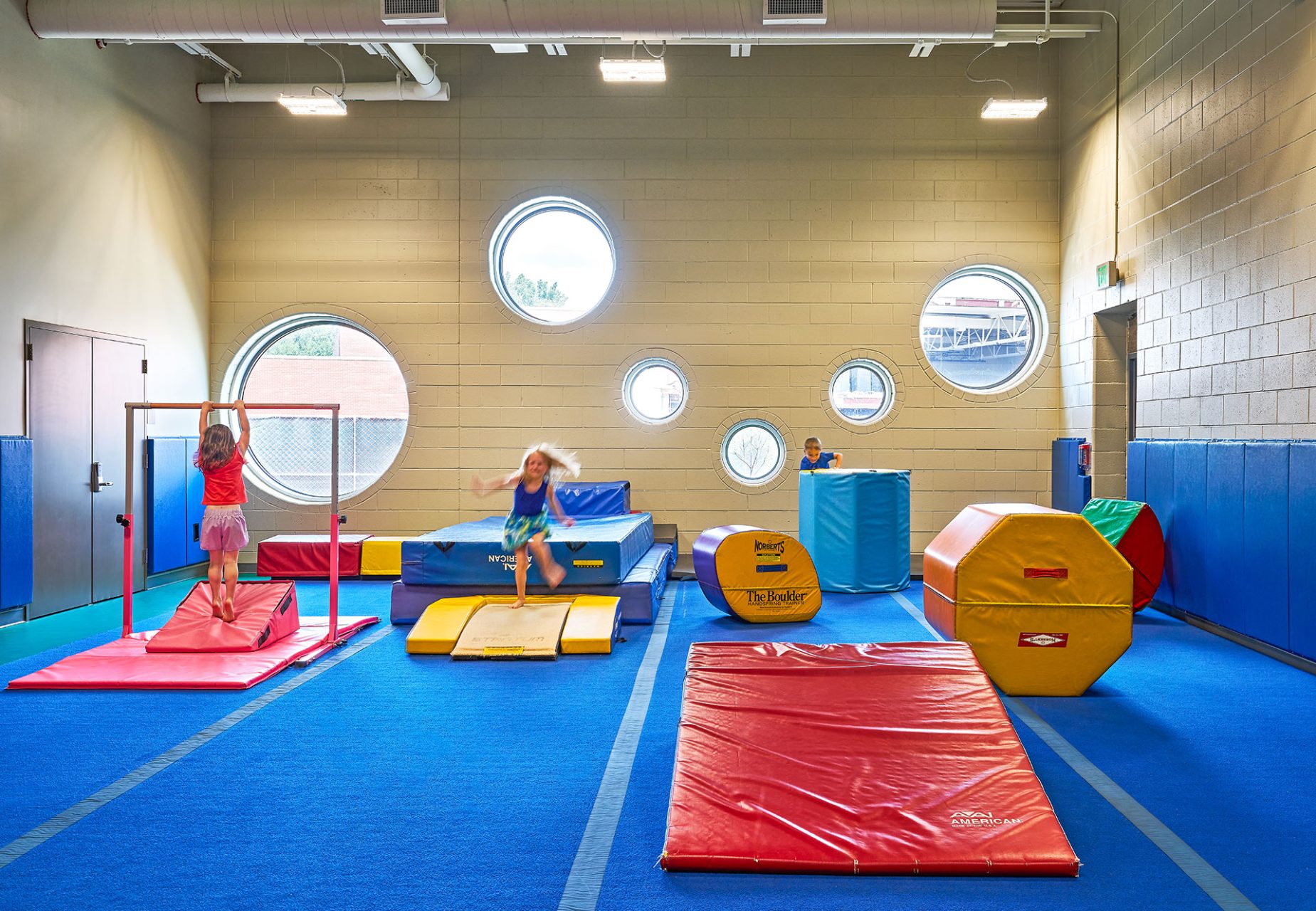 kids gymnastics area with padded play equipment, bars, and circular windows to the outside